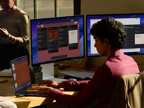 Un gruppo di persone sedute attorno a un tavolo in un ufficio. Una donna seduta alla scrivania che lavora su un MacBook con vari monitor. I colleghi e le colleghe sono riuniti intorno a lei a discutere.