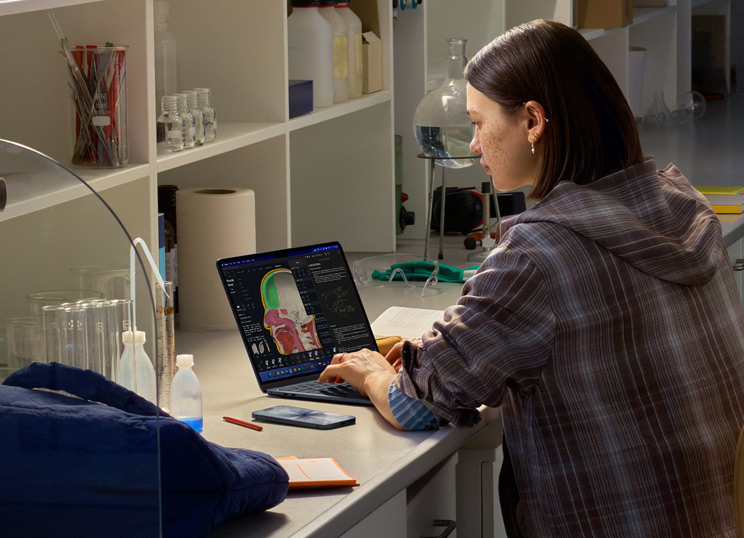 Une personne assise dans un laboratoire utilisant un MacBook dont l’écran affiche plusieurs apps.