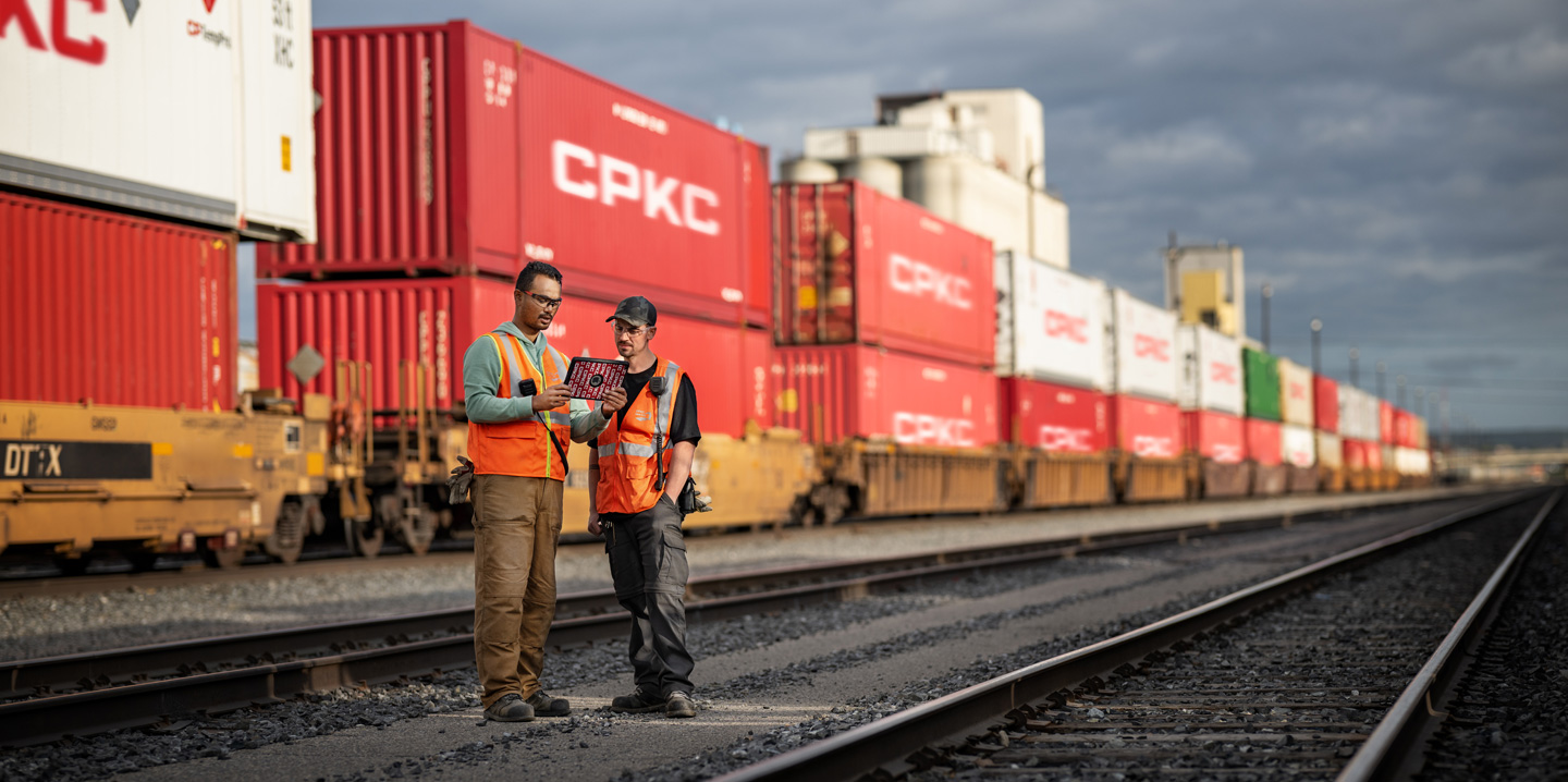 Duas pessoas da equipe da ferrovia consultam um iPad no pátio ferroviário, ao lado de um trem parado carregado de contêineres