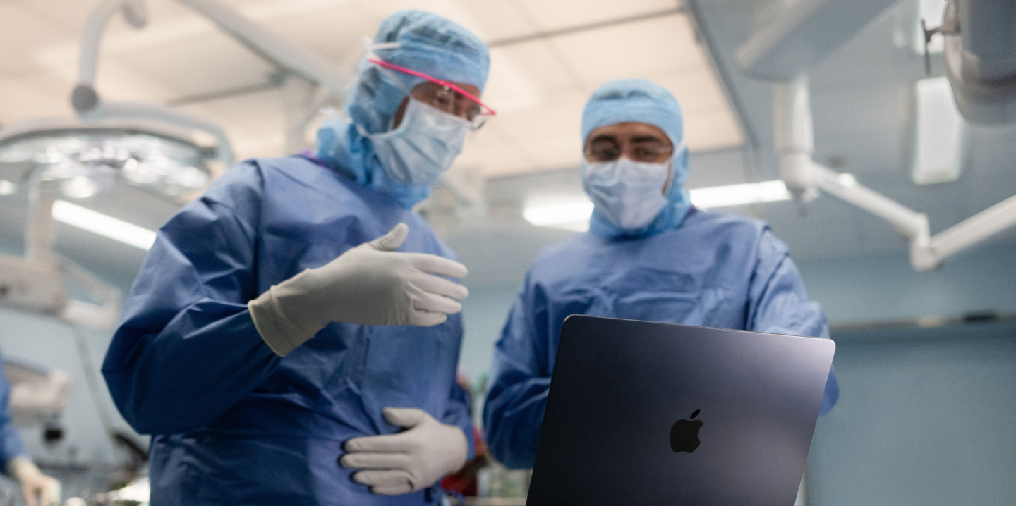Dois cirurgiões olham para um MacBook na sala de cirurgia, e um gesticula com a mão enquanto o outro observa