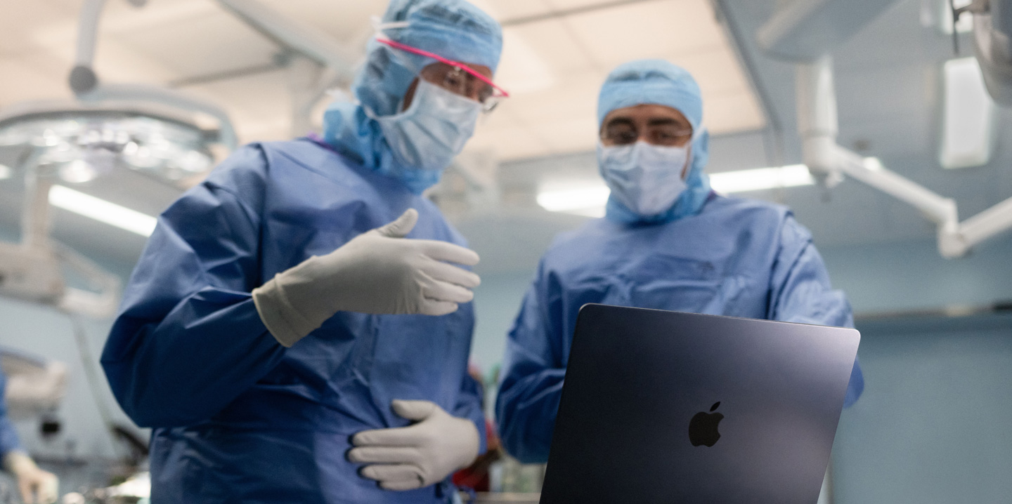 To kirurger på operationsstuen ser på en MacBook, og den ene gestikulerer med hånden, mens den anden ser på