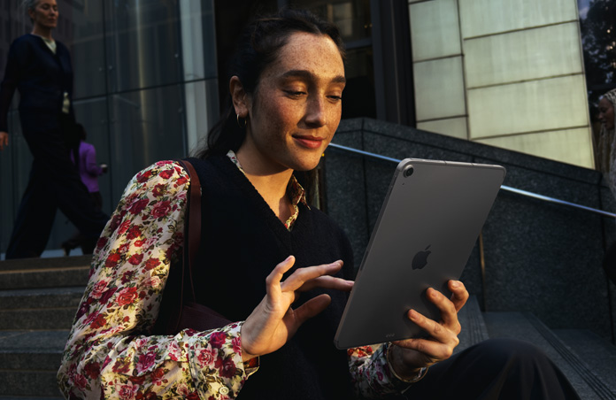 En person sidder på en trappe uden for en stor bygning og bruger en 11-tommers iPad Air i space grey med stabil Wi‑Fi-forbindelse under netværksskift